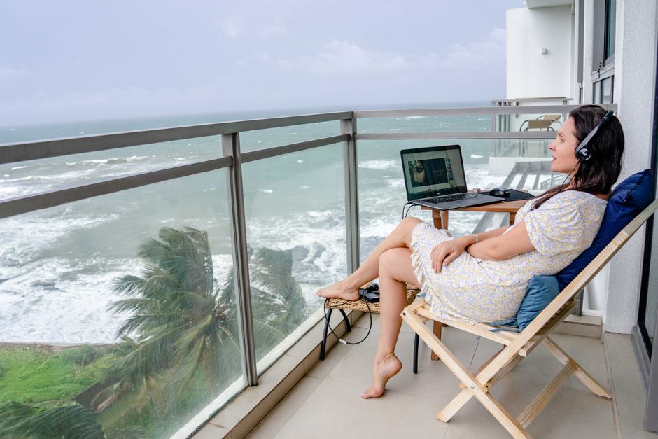 A woman wearing headphones relaxes on a balcony chair with a laptop, enjoying a seaside view.