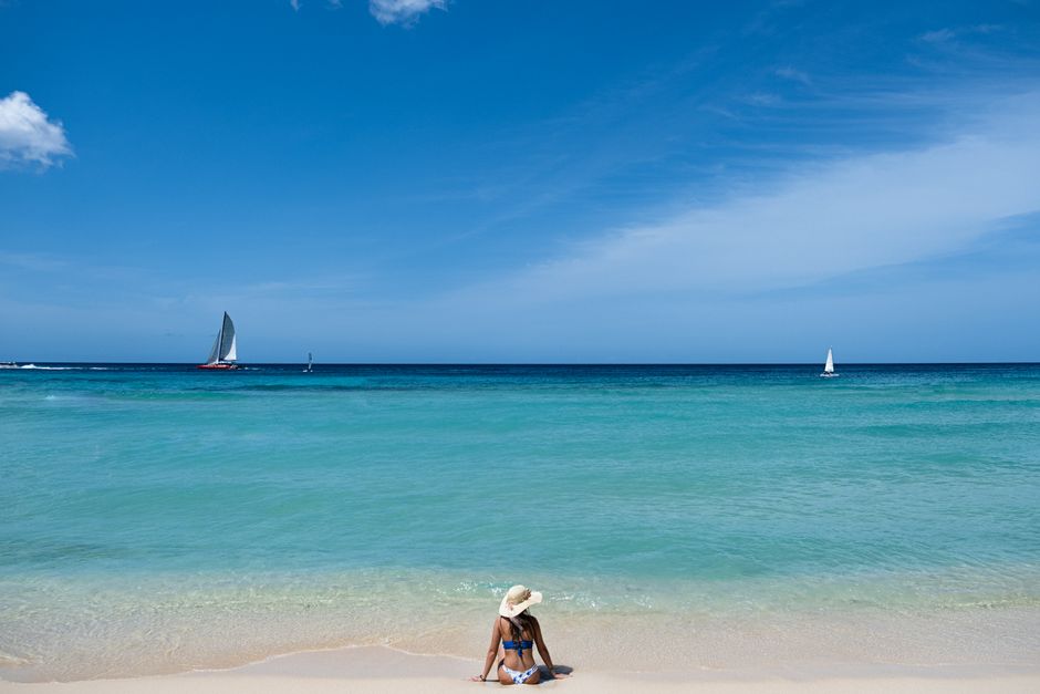 Woman in sun hat sitting on a tranquil beach with sailboats in the ocean.