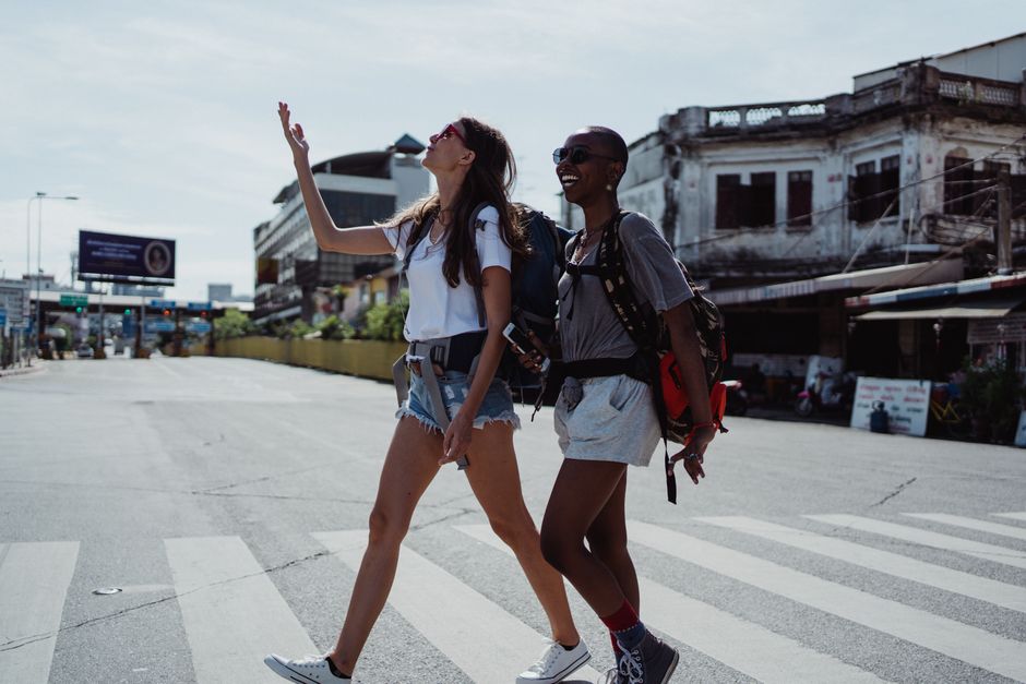 Two women with backpacks walk across a street, smiling joyfully under sunny skies.