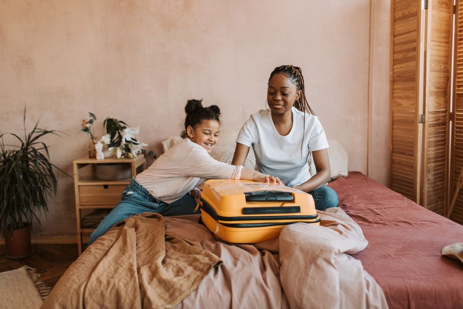 A mother and daughter share a joyful moment packing a suitcase on the bed.