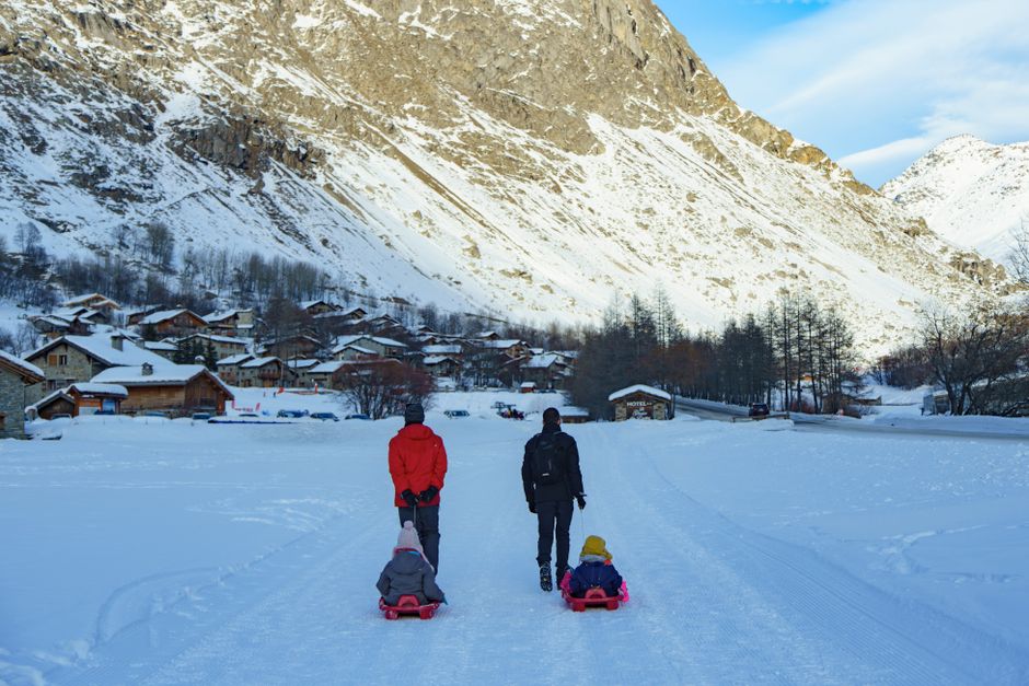 A family enjoys a winter day sledding along a snowy path in a picturesque village beneath snowy mountains.
