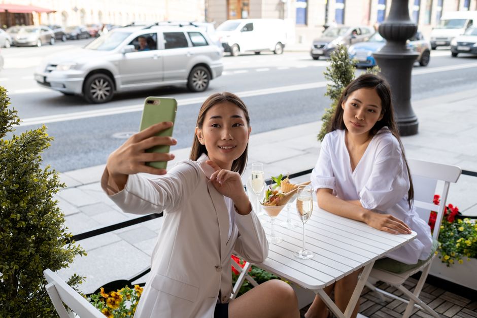 Two women taking a selfie at an outdoor cafe table on a sunny day.