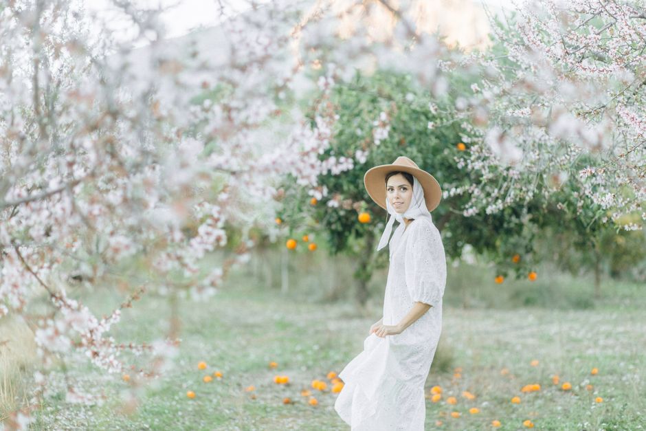 A woman in a white dress and hat stands gracefully in a blooming orchard.