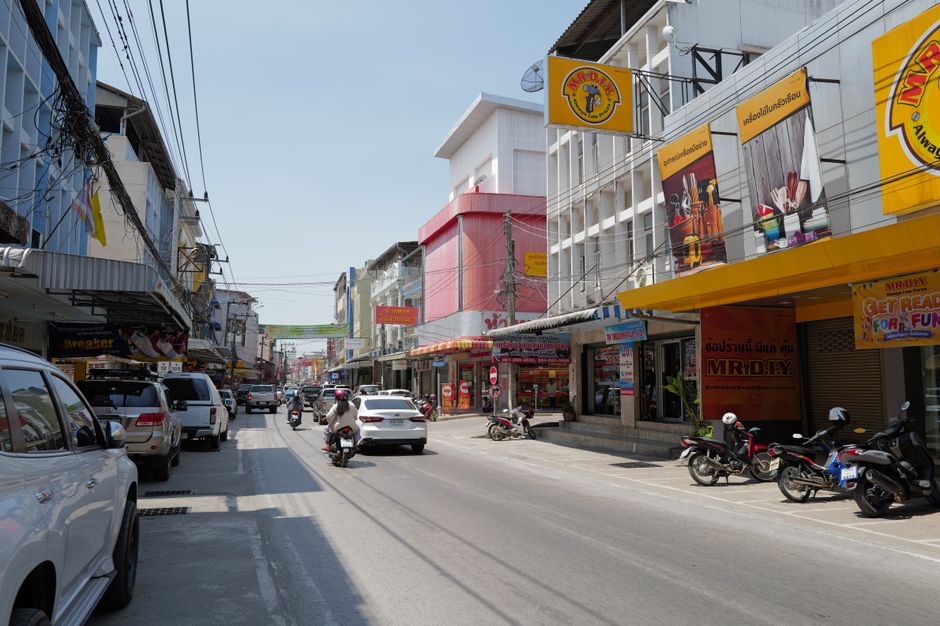 Lively Thai city street scene with cars, motorcycles, and colorful shop facades on a sunny day.