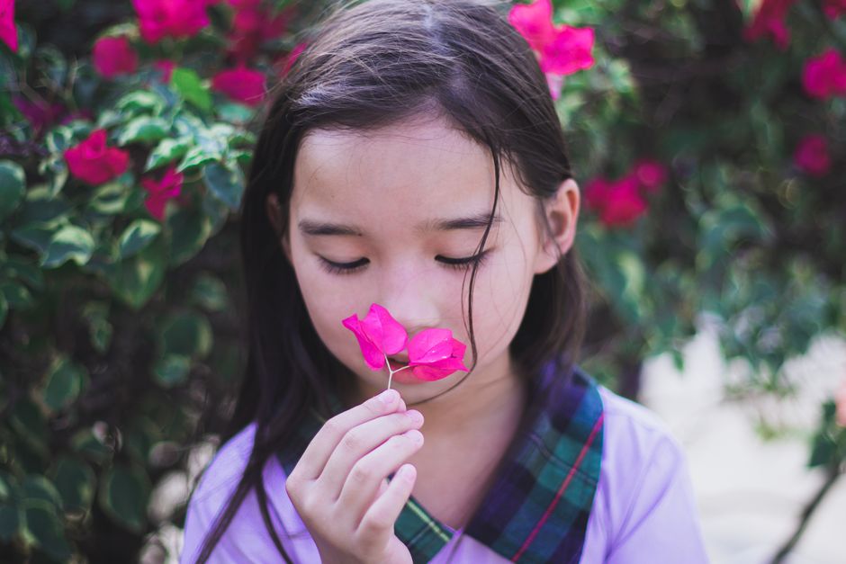 A young girl in a purple dress enjoying the scent of pink bougainvillea flowers in a vibrant garden setting.
