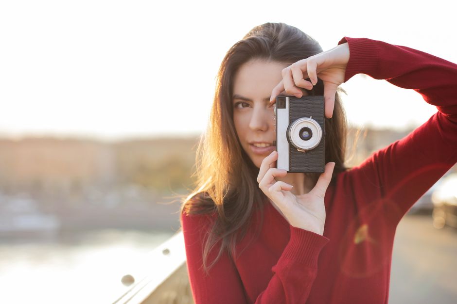 Brunette woman in red sweater posing with a vintage camera outdoors during sunset.